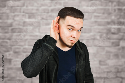 Young bearded man eavesdrops with the hand at ear on brick wall background
