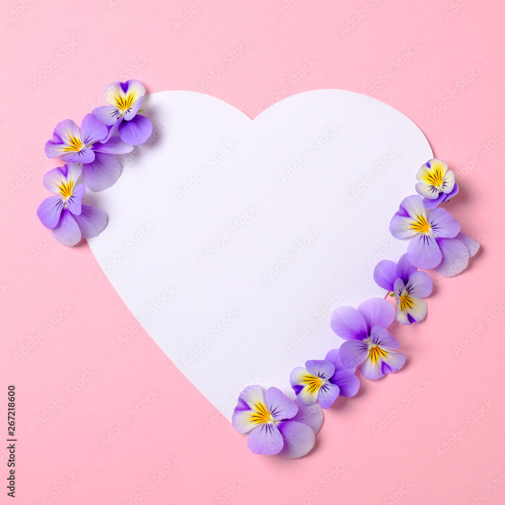 Heart-shaped blank paper card and wild violet flower petals on pastel pink background. Top view, floral frame, abstract design. Invitation, greeting card or an element for your design