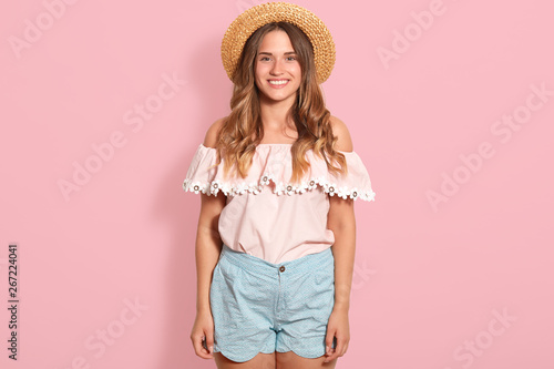 Happy girl posing in straw hat, rose summer blouse and blue short. Beautiful woman has charming smile, being in good mood, expresses happyness, looking directly at camera, standing isolated on pink.