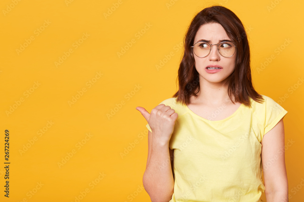 Good looking brunette female dressed in casual yellow t shirt and stylish eyeglasses, looking aside, showing direction with her thumb, looks astonished. Emotions concept. Copy space for advertisement. Good looking brunette female dressed in casual yellow t shirt and stylish eyeglasses, looking aside, showing direction with her thumb, looks astonished. Emotions concept. Copy space for advertisement.
