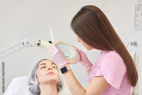 Long haired beautician standing near her client, touching magnifying glass, watching through it, exploring skin condition. Model in medical cap looks at beautician. People and procedures concept. photo