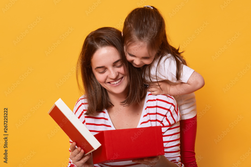 Family, child, holiday and Mother's Day concept. Smiling young woman in striped shirt sitting with her toddler daughter and opening red box, present for holiday, liitle girl in t shirt and trousers. - obrazy, fototapety, plakaty 