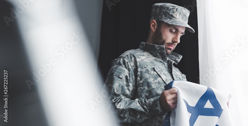 selective focus of pensive military man in uniform holding israel national flag while standing by window