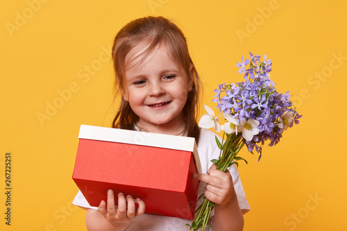 Portrait of beautiful little girl with red gift and bouquet of blue florets, preparing for Mother's Day, wants to congratulate her mommy, holds present in hands