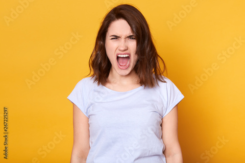 Screaming emotional angry woman isolated over yellow studio background. Half length portrait of young brunette female wearing white casual t shirt. Human emotions and facial expression concept.