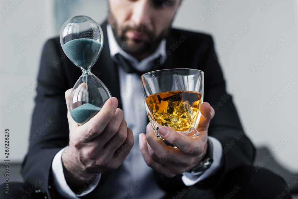 cropped view of depressed businessman holding hourglass and glass of whiskey cropped view of depressed businessman holding hourglass and glass of whiskey