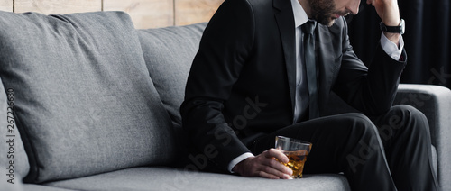 panoramic shot of depressed man sitting on sofa with glass of whiskey