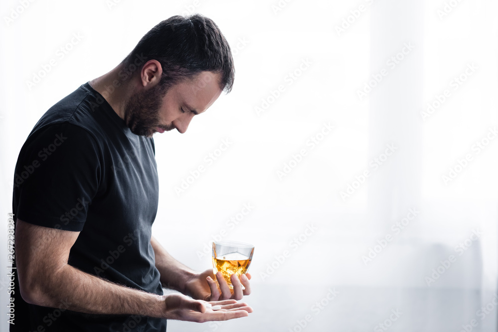 depressed man in black t-shirt standing with glass of whiskey and handful of pills depressed man in black t-shirt standing with glass of whiskey and handful of pills