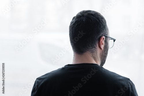 Fotografia back view of adult depressive man in black t-shirt looking out window at home