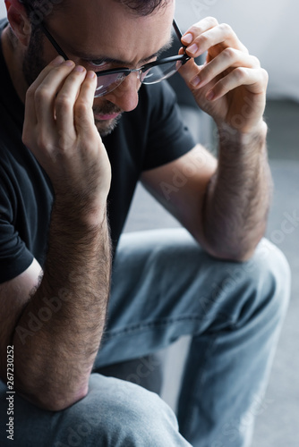 upset man touching glasses while sitting on sofa at home