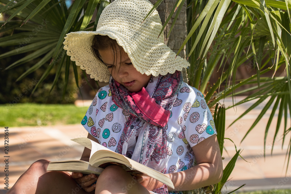 Dziewczynka w słomkowym kapeluszu czyta książkę w cieniu palmy - obrazy, fototapety, plakaty 