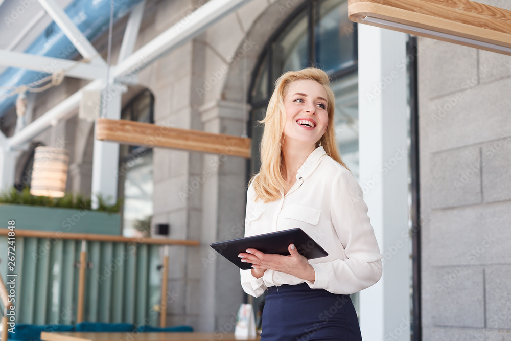 Fototapeta premium Positive beautiful blonde - the manager of large financial company holds tablet and looks somewhere during meeting in a large beautiful office.