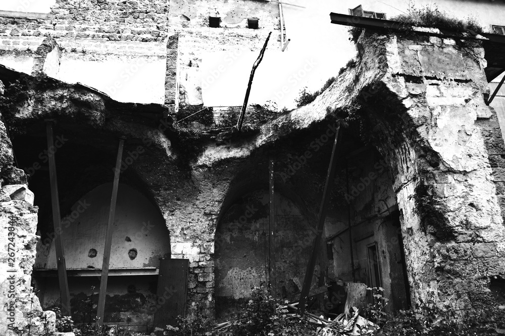 Abandoned ruined house near residential building. Broken stone wall and arches supported by rusty metal props. Collapse danger concepts. Safety problem background. Black white photo.