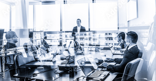 Successful team leader and business owner leading informal in-house business meeting. Businessman working on laptop in foreground. Business and entrepreneurship concept. Blue toned grayscale.