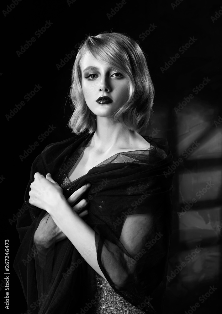 Cute face blond girl with vintage style hairstyle, wearing a golden sparkling dress and black veil on her shoulders stands on the background of a wall with window shadow