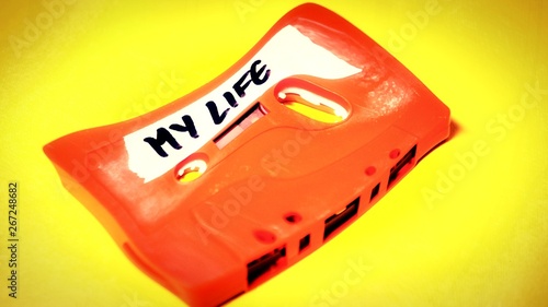 Heavy wave distortion fx: a vintage cassette tape (obsolete music technology), orange on a yellow surface, angled shot, carrying a label with the handwritten text My life.