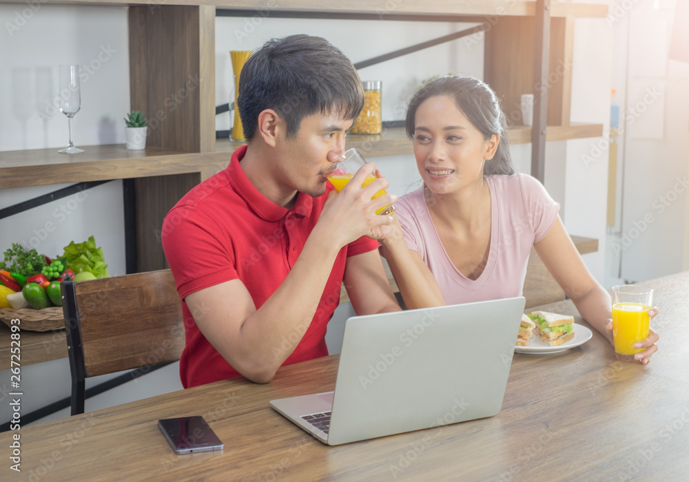 Asian young couple, sitting at the dining table. they are happily. smiling drinking orange juice. And have a sandwich in plate with laptop computer and mobile phone on the table placed in front. Asian young couple, sitting at the dining table. they are happily. smiling drinking orange juice. And have a sandwich in plate with laptop computer and mobile phone on the table placed in front.