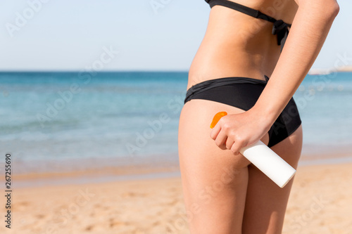 Young beautiful girl in black bikini is applying sunblock from the spray on her buttocks at the sea background