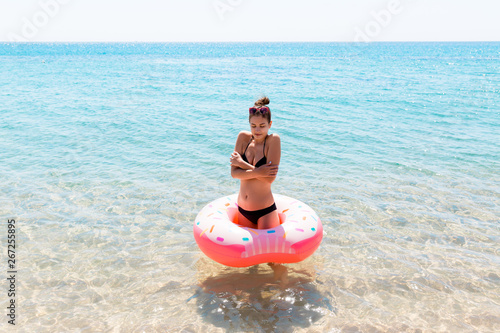 Woman swimming with inflatable donut in cold shivering sad crossed arms black bikini swimsuit standing in sea water. Summer holidays and vacation concept