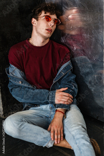 Stylish young fashion model man in bright red sunglasses and denim casual style sitting on floor and posing near metallic door and looking up. indoor, studio shot.