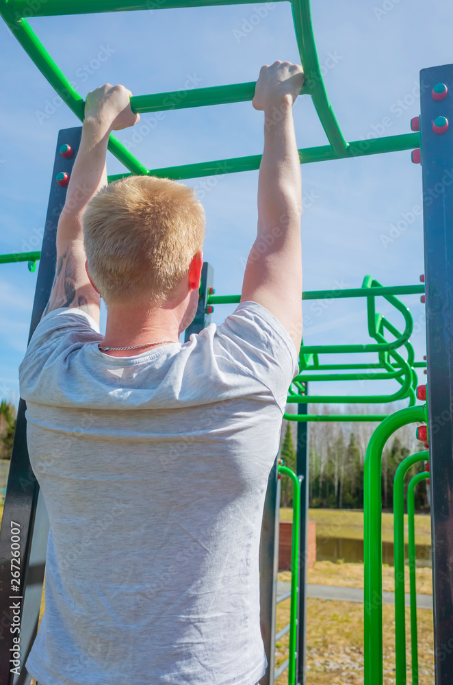 Fototapeta premium The concept of a healthy lifestyle, sports in nature, physical culture. A blond man pulls himself up on a crossbar on a street stadium. Summer sunny day.