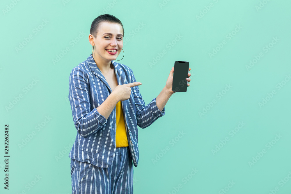 Happy beautiful with short hair young woman in striped suit standing, holding phone and pointing finger to it, toothy smile and looking at camera. Indoor, isolated, studio shot, green background Happy beautiful with short hair young woman in striped suit standing, holding phone and pointing finger to it, toothy smile and looking at camera. Indoor, isolated, studio shot, green background