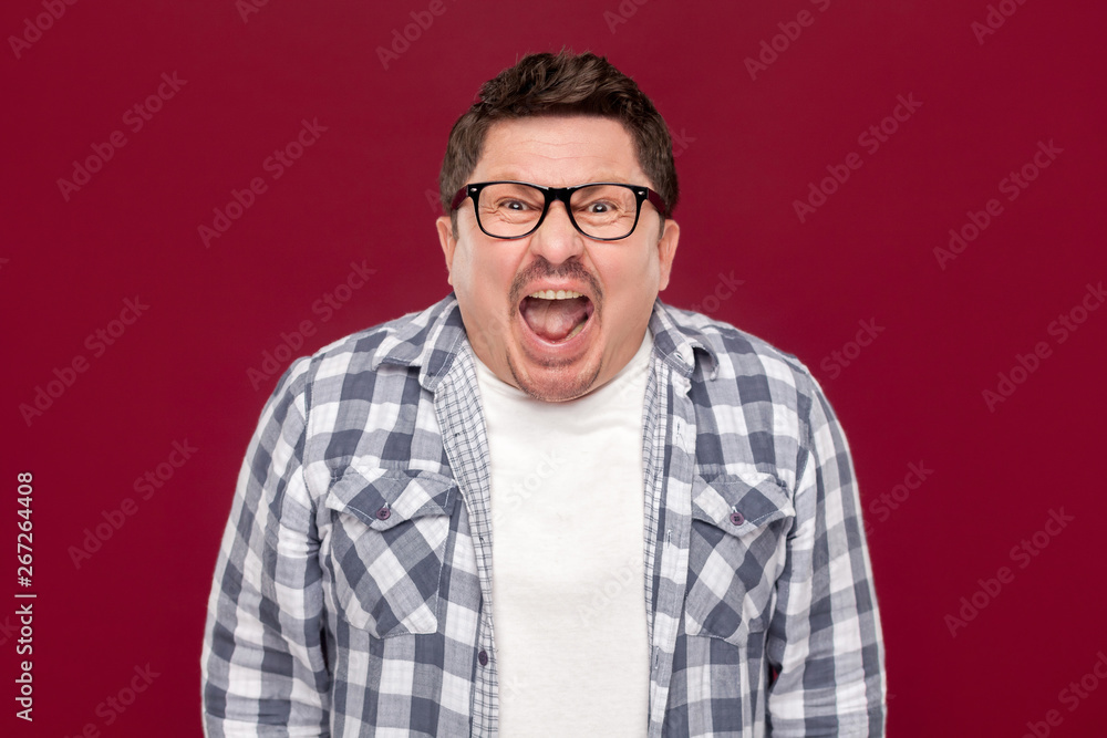 Portrait of angry handsome middle aged business man in casual checkered shirt and eyeglasses standing, looking and screaming at camera. indoor studio shot, isolated on dark red background. Portrait of angry handsome middle aged business man in casual checkered shirt and eyeglasses standing, looking and screaming at camera. indoor studio shot, isolated on dark red background.