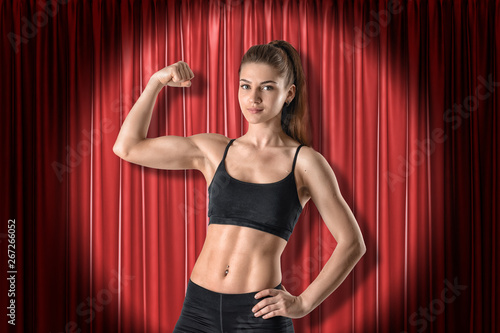Young athletic girl in black sport clothing showing biceps on red stage curtains background