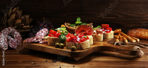 Traditional italian antipasto bruschetta appetizer with cherry tomatoes, cream cheese, basil leaves and balsamic vinegar on cutting board with prosciutto, salami, cheese