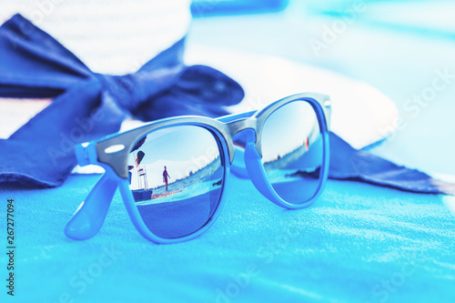 White straw summer hat with blue sunglasses at seaside with a blurred person reflection. Travel and marine vacation concept over a blue background with copy space