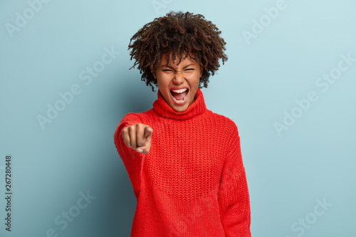 Strict emotional annoyed woman being furious, points at camera with anger and digust and blames you, screams loudly, wears red knitted sweater, isolated over blue background. Negative emotions