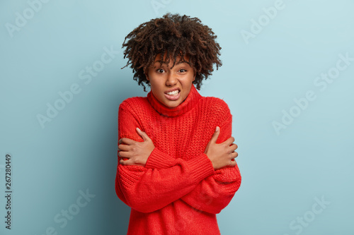 Waist up shot of dissatisfied dark skinned woman crosses hands over chest, feels scared, trembles from fear, frowns face, wears red warm sweater, isolated over blue background. Its chilling there