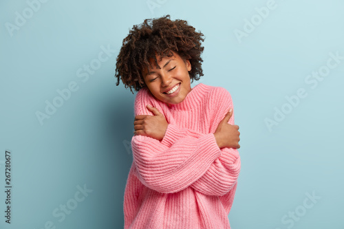 Love yourself concept. Photo of lovely smiling woman embraces herself  has high self esteem  closes eyes from enjoyment  likes her new comfortable soft pink sweater  tilts head  stands indoor