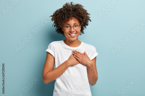 Indoor shot of happy dark skinned lady swears or promises something, holds hands on chest, tells truth, being honest, looks at camera friendly, has charming smile, wears casual white t shirt, glasses photo