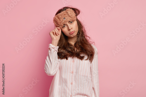 Displeased European woman wears bear sleep mask, pyjamas, tilts head, purses ...