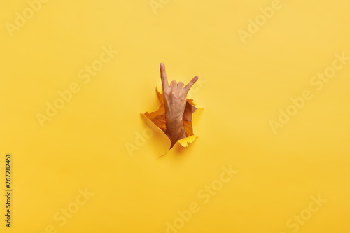 Unrecognizable man makes rock n roll gesture through ripped hole in yellow paper. Male demonstrates horn sign with hand stretched in gap slot of paper. Body language concept. Colored background