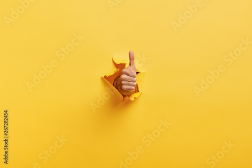 Image of unrecognizable man makes thumb up gesture, demonstrates approval or agreement, gestures through torn paper wall yellow background. Body language concept. Hand sign. Hole in wall. Like gesture photo