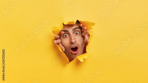 Stupefied dark eyed European man tears yellow paper, stares through hall with scared expression, notices something terrified, has widely opened eyes and mouth. Yellow background. Omg concept
