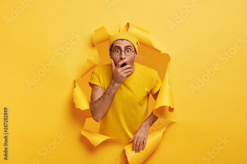 Photo of surprised scared man breaks through color paper wall, covers mouth, has stupefied expression, can not believe his eyes, wears stylish headgear, eyewear and t shirt in one tone with background photo