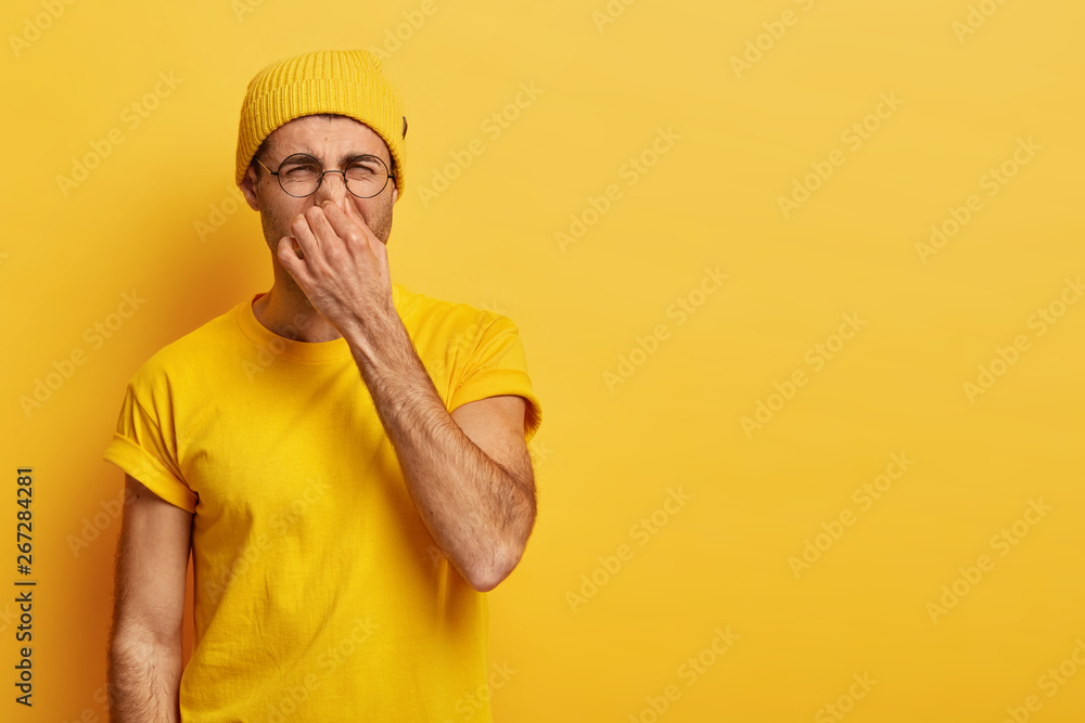 Disgusted young hipster pinches nose with fingers, looks in disgust as smells something stink, wears spectacles, yellow hat, t shirt, stands in stuido with blank copy space. Fie, unpleasant smell Disgusted young hipster pinches nose with fingers, looks in disgust as smells something stink, wears spectacles, yellow hat, t shirt, stands in stuido with blank copy space. Fie, unpleasant smell
