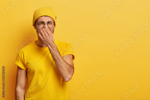 Disgusted young hipster pinches nose with fingers, looks in disgust as smells something stink, wears spectacles, yellow hat, t shirt, stands in stuido with blank copy space. Fie, unpleasant smell