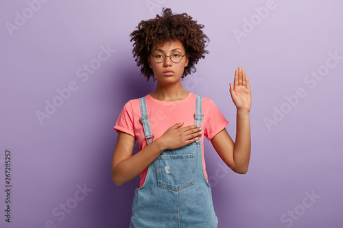Confident serious woman with curly hair makes sincere promise or oath, keeps one hand on heart, solemnly swears, raises palm, demonstrates loyalty gesture being honest poses against purple background. photo