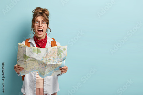 Photo of frustrated woman traveler lost in city, looks depressed at paper map, shouts from despair, has tour on vacation, carries backpack, wears big round spectacles models indoor copy space for text photo