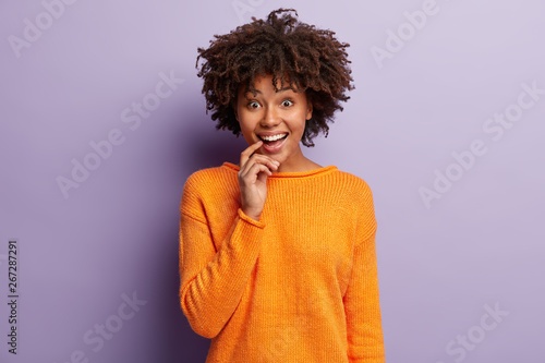 Studio shot of glad mirthful woman keeps fore finger on lips  smiles delighted and upbeat at camera  wears orange jumper  shows white teeth  isolated over purple background. Positive emotions