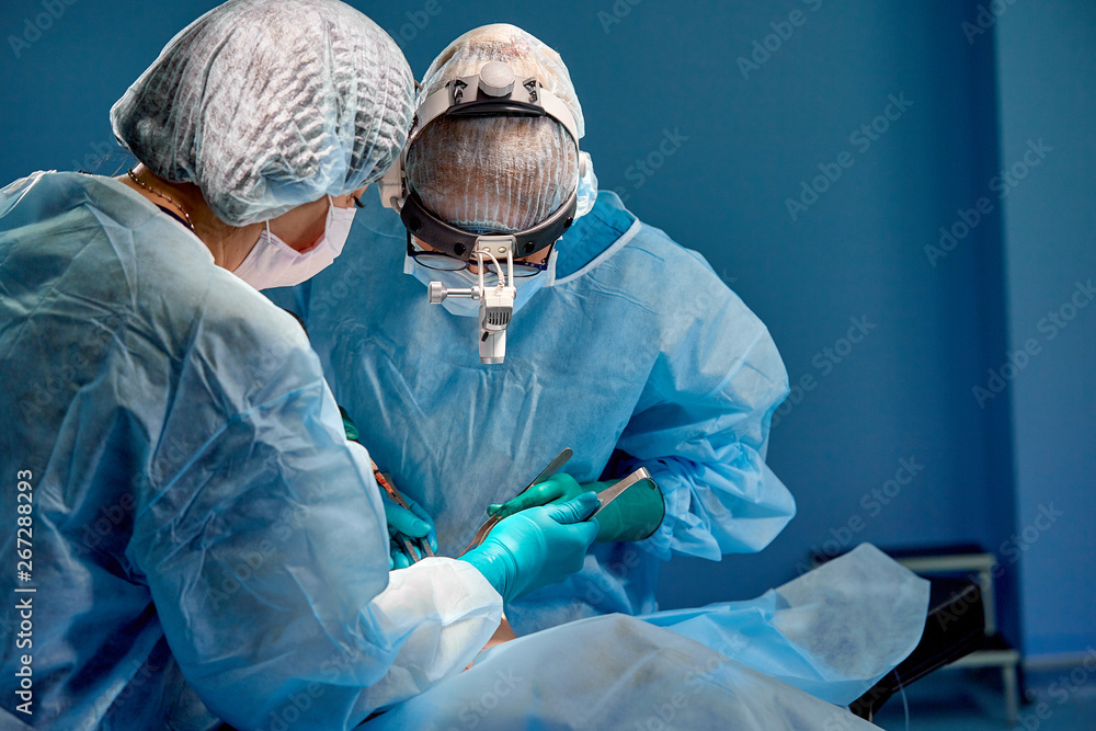 Surgeon and his assistant performing cosmetic surgery in hospital operating room. Surgeon in mask wearing loupes during medical procadure. Surgeon and his assistant performing cosmetic surgery in hospital operating room. Surgeon in mask wearing loupes during medical procadure.