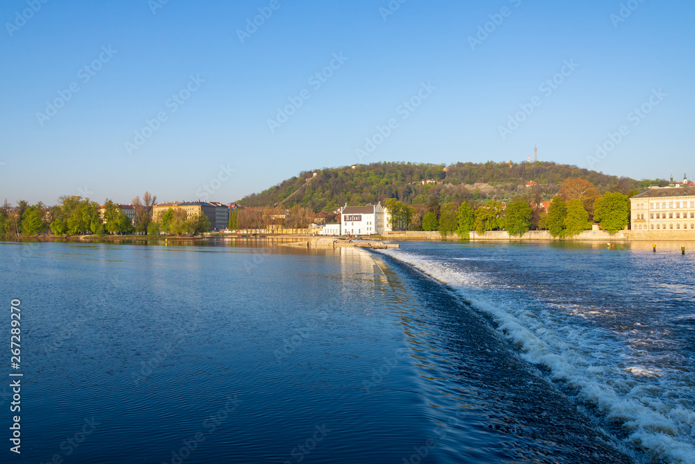 Outdoor sunny scenery of Vltava river with Museum Kampa on riverside, weir on the river, and Petrin Tower on the hill in Prague, Czech Republic. 
