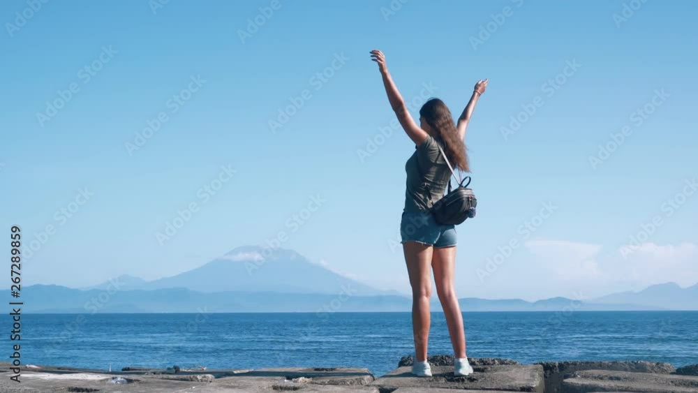 Girl traveler with backpack spreads her arms wide, enjoys view of ocean, volcano Girl traveler with backpack spreads her arms wide, enjoys view of ocean, volcano