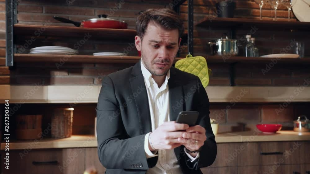 Handsome emotional Caucasian man in formal suit sitting at the kitchen table and actively messaging via smartphone, reacting negatively on received sms. Bad mood, bad news, messaging app Handsome emotional Caucasian man in formal suit sitting at the kitchen table and actively messaging via smartphone, reacting negatively on received sms. Bad mood, bad news, messaging app