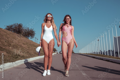 Two girlfriends women walk in the summer in city in white bodysuit. Emotions of joy, fun rest, walk on the weekend. Concept style fashion, lifestyle of young people. Long hair tanned figure.