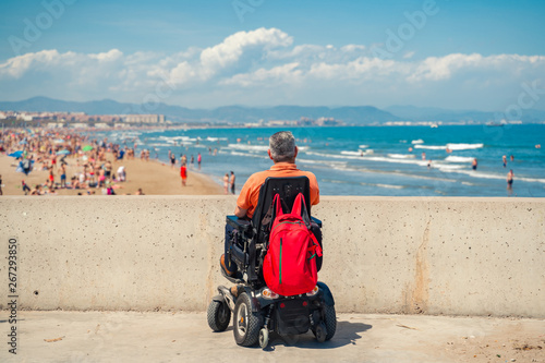 Lonely man on electric wheelchair looking  at the beach. Disabled person at the beach.
